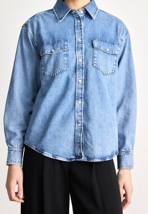 Person wearing light blue denim button-up shirt with two chest pockets and black pants, shown from neck to hips.