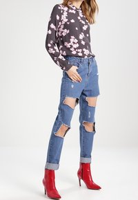 Blouse noire à motif floral avec manches longues, associée à un jean bleu déchiré avec de grandes déchirures aux genoux, et des bottines rouges à talons hauts.