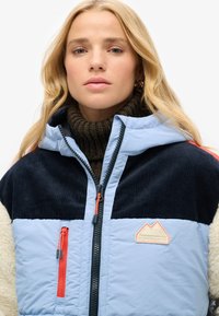 Giacca imbottita di colore azzurro chiaro con parte superiore in corduroy blu navy, lati in pile crema e tasca con zip; presenta una cerniera arancione e una patch a forma di montagna.