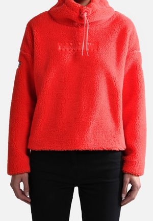 Personne portant un pull polaire rouge vif à demi-fermeture éclair et capuche à cordon, associée à un pantalon noir, debout les mains détendues.