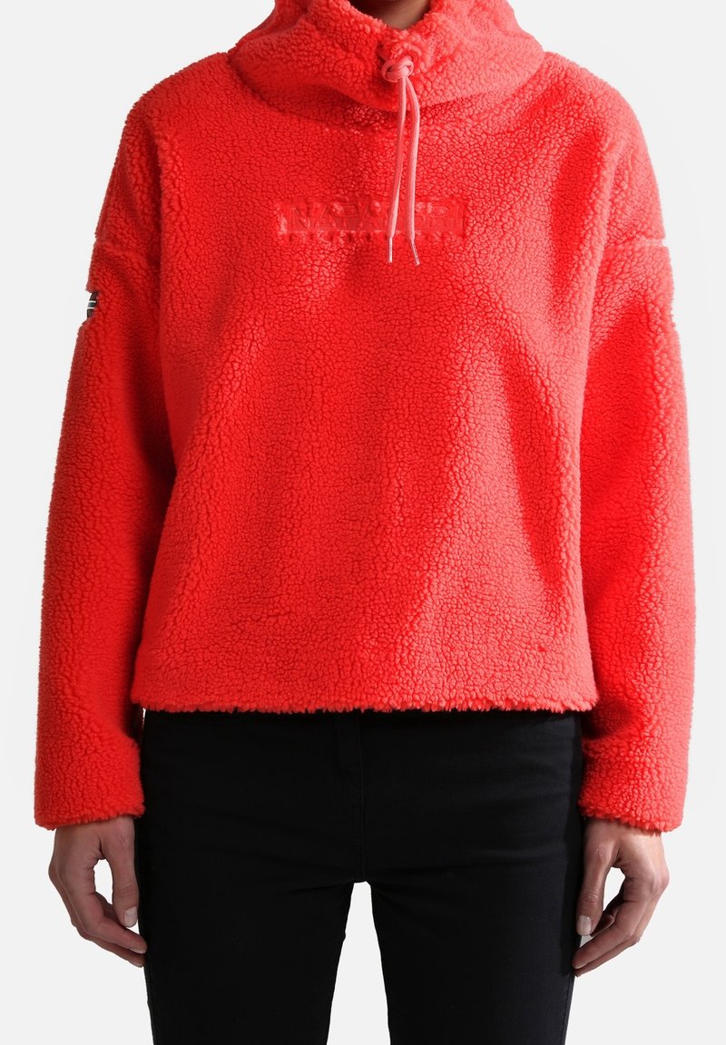 Personne portant un pull polaire rouge vif à demi-fermeture éclair et capuche à cordon, associée à un pantalon noir, debout les mains détendues.