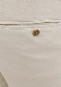 Gros plan sur un tissu beige avec une poche cousue et un seul bouton marron fixé près de l'ouverture de la poche.