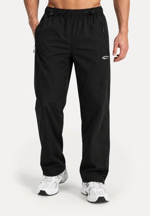 Hombre que lleva pantalones casuales negros con cintura elástica y bolsillos con cremallera, combinado con zapatos deportivos blancos, de pie con las manos cerca de las caderas.