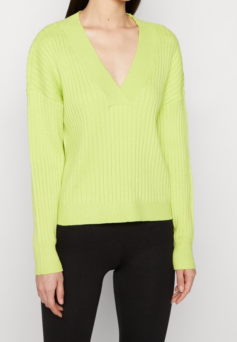 Femme portant un pull à col en V côtelé vert citron à manches longues et un pantalon noir ajusté, sur un fond blanc uni.