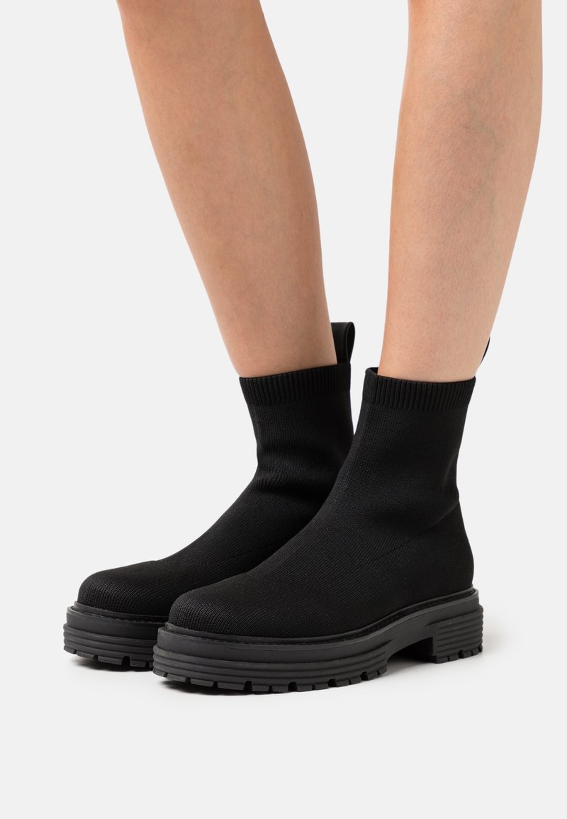 Botas negras hasta el tobillo con parte superior de punto, que cuentan con una suela de goma gruesa y una superficie texturizada. El diseño incluye una lengüeta en la parte trasera.