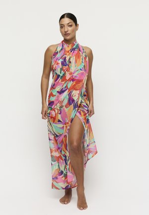 Vrouw in een meerkleurige halterjurk met bloemenprint en een hoge split, blootsvoets staand tegen een effen lichte achtergrond.