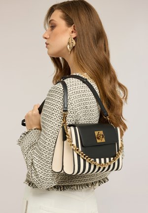 Mujer con cabello ondulado lleva pendiente de hoja de oro y chaqueta de tweed, llevando un bolso de mano a rayas negras y blancas con cadena y cierre dorado en el hombro.