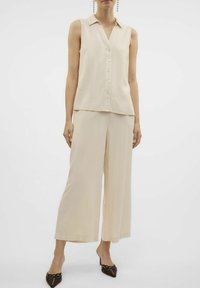 Beige ärmlös blus med knappar framtill, kombinerad med vida byxor i matchande färg. Har lätt material och minimalistisk design.