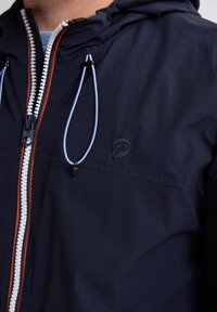 Marineblauwe jas met een ritssluiting aan de voorkant, voorzien van een witte rits en oranje accenten. Verstelbare trekkoorden bij de capuchon en een gestructureerde stof.