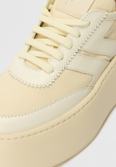 Beige sneaker featuring a mix of textured canvas and smooth leather, rounded toe, and flat laces, with a chunky rubber sole.