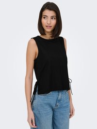 Woman with straight dark hair wearing a sleeveless black top with side ties and blue jeans, looking to her left against a plain light background.