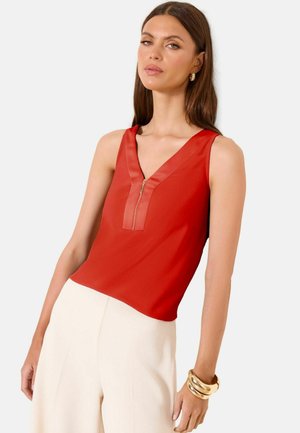 Femme portant un top rouge sans manches avec fermeture éclair à l'avant, associé à un pantalon blanc taille haute et des boucles d'oreilles créoles dorées ainsi que des bracelets.