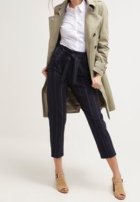 En beige trenchcoat over en hvid skjorte med knapper, parret med marineblå stribede kapri-bukser og brune hæle, der viser en skræddersyet pasform.