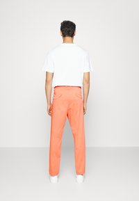Herrenweite, leuchtend orangefarbene Hose mit Taschen, kombiniert mit einem schlichten weißen T-Shirt und weißen Sneakers. Unifarbener Stoff, ohne sichtbare Muster.