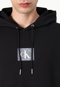 Czarna bluza z kapturem z miękkiego materiału, z prostokątną grafiką z napisem "Calvin Klein" oraz "New York City", z sznurkami zakończonymi srebrem.