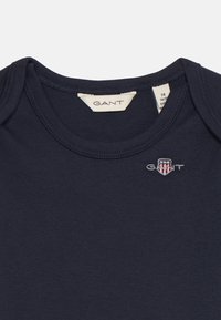 T-shirt en coton bleu marine avec un col rond, arborant un petit logo GANT brodé et une étiquette avec des informations sur la taille et l'entretien.
