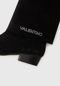 Botte de cheville en suède noir avec un talon bloc. Présente "VALENTINO" en cristaux cloutés sur le côté extérieur. Texture lisse, design épuré.