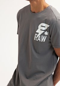 Šedé bavlněné tričko s kulatým výstřihem a krátkými rukávy. Na levé straně hrudi výrazné bílé logo "RAW". Hladká textura.