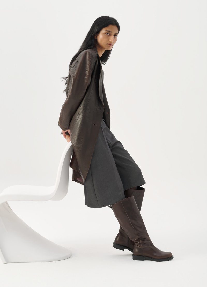 Veste en cuir marron sur short gris ajusté, associée à des bottes marron jusqu'aux genoux, sur fond blanc uni.