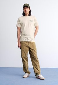 Camiseta de algodón beige con pequeño logo verde, combinada con pantalones de pana marrón claro y zapatillas blancas con rayas rojas. Gorro verde.