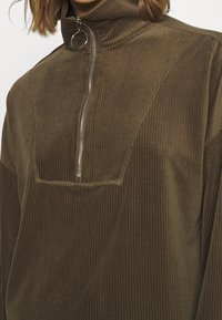 Sudadera de pana marrón con cuello alto, media cremallera y un gran bolsillo frontal. Presenta una textura vertical y detalles de costura en contraste.