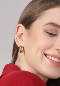 Ohrringe aus vergoldetem Metall in Form von Creolen, die einen auffälligen dunklen Edelstein und strukturierte Akzente zeigen und mit einer glatten, polierten Oberfläche glänzen.