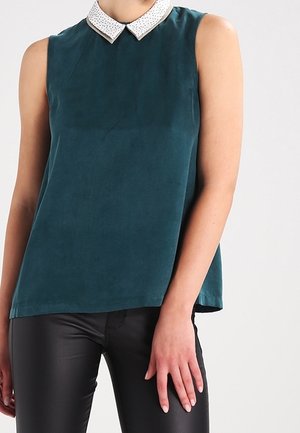 Ærmeløs mørk teal top med hvid mønstret Peter Pan-krave båret med sorte tætsiddende bukser på en person, der står.