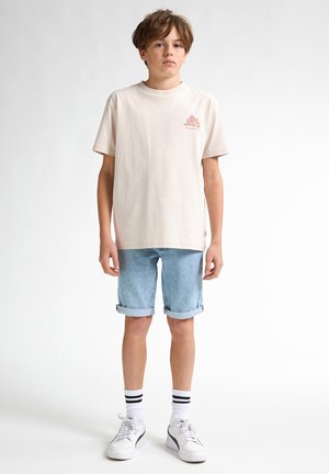 T-shirt di cotone beige chiaro con una piccola grafica rosa e pantaloni shorts di denim corti e arrotolati. Sneakers bianche con dettagli neri e calze a righe.