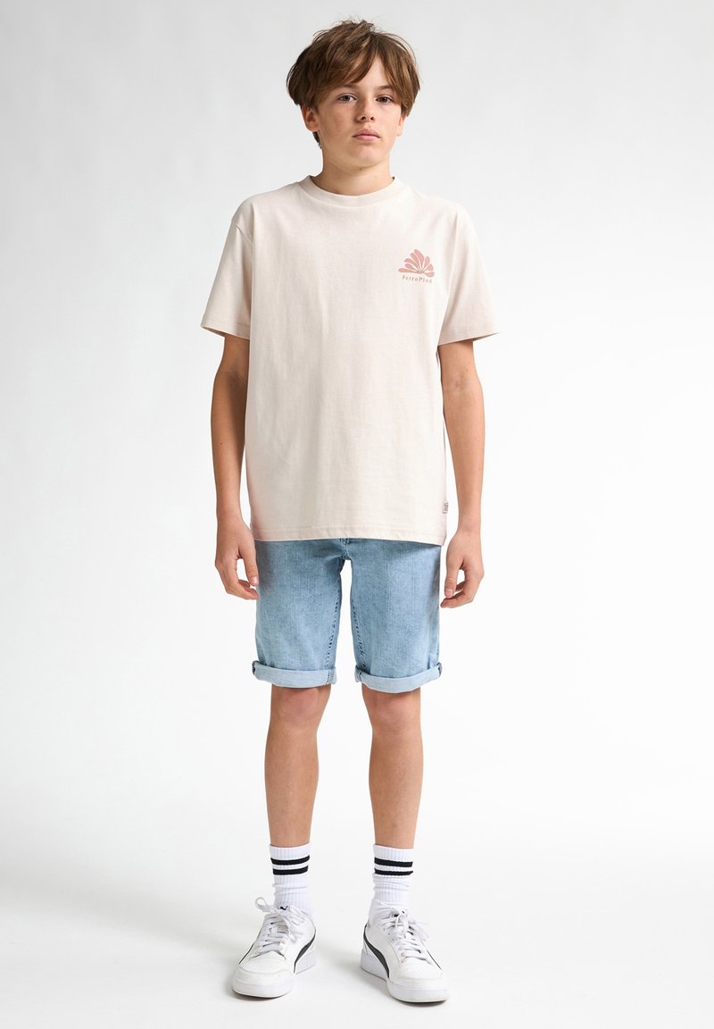 Licht beige katoenen T-shirt met een kleine roze grafiek en opgerolde korte denim shorts. Witte sneakers met zwarte accenten en gestreepte sokken.