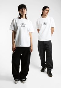White cotton T-shirts with a central black graphic, paired with loose black pants; one wearing sneakers, the other in black footwear.
