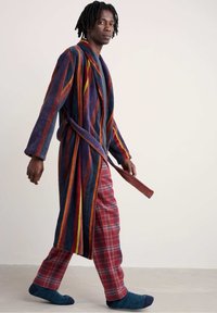 Gestreepte fleece badjas met blauwe, rode en gele tinten, vastgebonden in de taille, gecombineerd met rode geruite broeken en blauwe sokken, op een neutrale achtergrond.