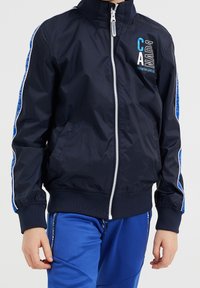 Lapsi, joka on pukeutunut tummansiniseen vetoketjulliseen takkiin, jossa on teksti "KANADA", ja sinisiin urheiluhousuihin, seisoo tasaisen taustan edessä.