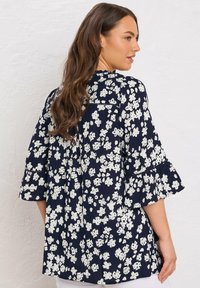 Vrouw met lang bruin haar, gekleed in een marineblauwe blouse met een wit bloemenpatroon en uitlopende mouwen, staande tegen een witte textuurwand.