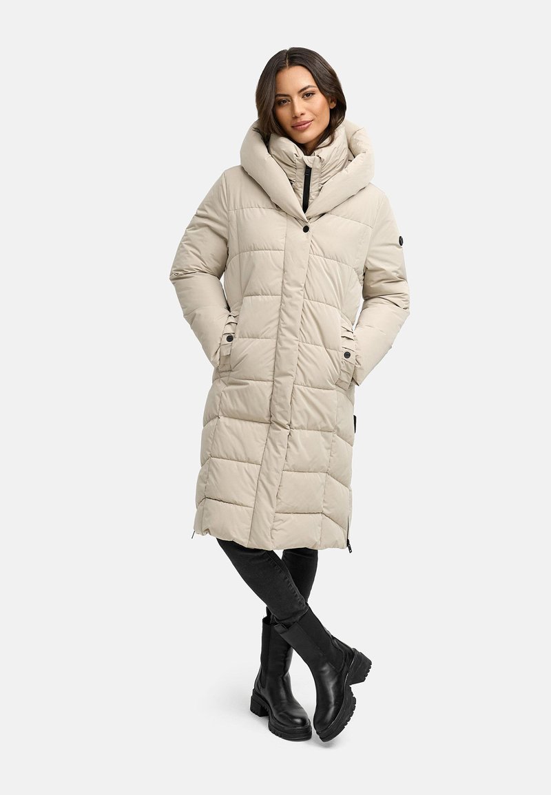 Beige, gepolsterter langer Mantel mit einer großen Kapuze, ausgestattet mit einem Knopfverschluss und seitlichen Taschen. Kombiniert mit schwarzen Hosen und Stiefeln, vor einem schlichten Hintergrund.