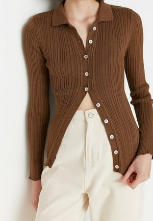 Cardigan côtelé marron avec un col, orné de boutons blancs sur le devant et présentant une silhouette ajustée. Assorti à un pantalon de couleur claire.