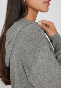 Sudadera gris con capucha, de tejido texturizado y puños acanalados. Vista cercana que muestra los detalles de las costuras y el ajuste alrededor del hombro.