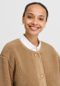 Beige fleecejacka med en mjuk, texturerad yta, med en rund krage och två bruna knappar. Bärs över en vit skjorta med krage.