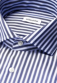 Primo piano di un colletto e pulsanti di una camicia a righe verticali blu e bianca con un'etichetta del marchio "seidensticker" all'interno del colletto.