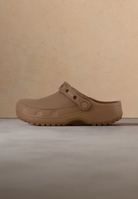 Sabot marron clair à enfiler avec semelle texturée et bride de talon réglable, sur fond de mur beige et sol gris.