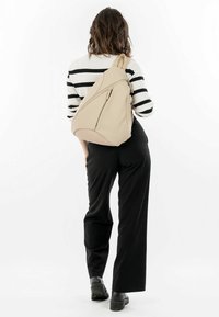 Beige ryggsäck med en slank, ergonomisk design, försedd med en framficka med dragkedja och en mjuk yta, som bärs över en svart outfit.