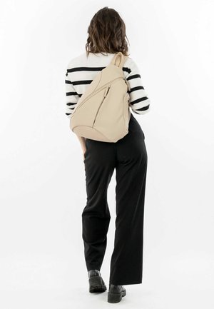 Sac à dos beige au design élégant et ergonomique, avec une poche zippée à l'avant et une texture lisse, porté avec une tenue noire.