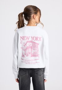 Nuori henkilö, jolla on ruskea poninhäntä, pukeutunut vaaleanharmaaseen huppariin, jossa on vaaleanpunainen New York ja Golden Bagel -myymälän graafinen kuvasto selässä.