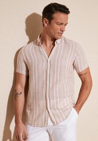 Camisa de botón de manga corta beige con rayas verticales blancas, hecha de tela ligera, con un cuello casual y un corte holgado.