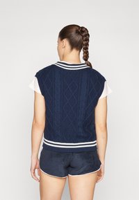 Gilet en maille bleu marine avec motif torsadé, col et ourlet côtelés rayés blancs, associé à un short sombre.