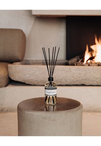 Diffuseur de parfum avec une bouteille en verre clair, des tiges noires et une étiquette indiquant "Courage des Bois." Posé sur une table ronde en béton.