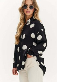 Czarna, długosleeve koszula zapinana na guziki z dużymi białymi kropkami; gładki materiał, luźny krój i boczne rozcięcia dla większej swobody ruchów.