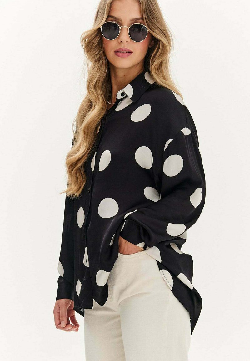 Czarna, długosleeve koszula zapinana na guziki z dużymi białymi kropkami; gładki materiał, luźny krój i boczne rozcięcia dla większej swobody ruchów.