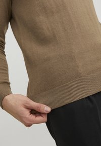 Pull en maille côtelée de couleur marron clair avec une coupe ajustée. Gros plan montrant la texture et une main saisissant l'ourlet. Associé à un pantalon foncé.