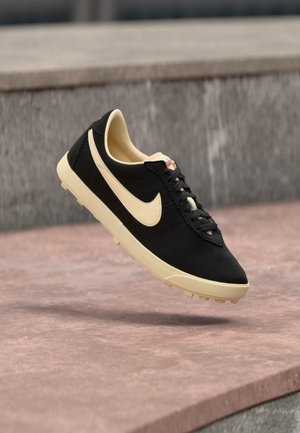 Black and beige Nike sneaker with laces, floating above a pinkish stone surface and gray concrete background.