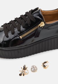 Baskets plateforme en cuir verni noir avec fermeture éclair dorée et trois pins décoratifs représentant une abeille, une pierre précieuse en forme de cœur et une coccinelle.